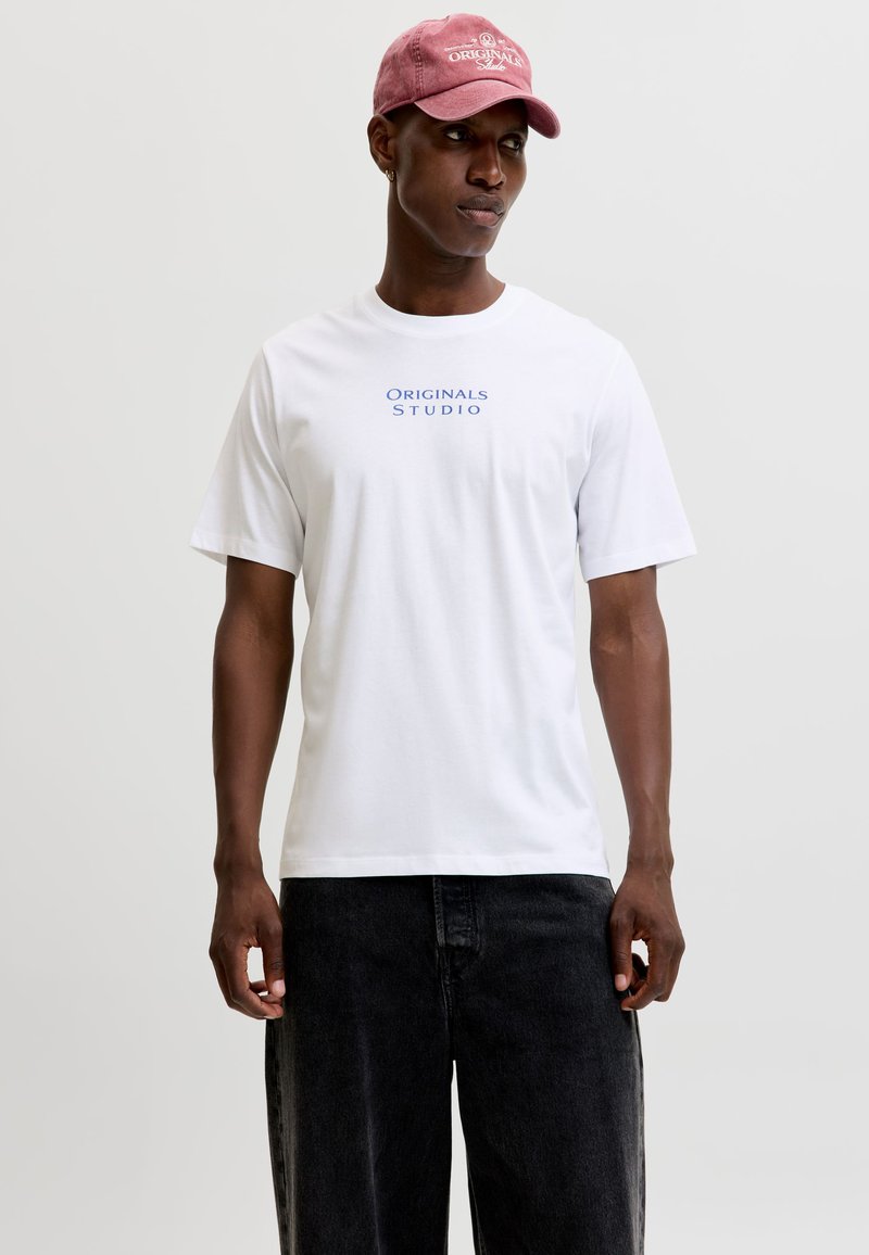 Homme portant un t-shirt blanc avec le texte "Originals Studio", un jean foncé et une casquette de baseball rouge délavée, debout et regardant sur le côté.