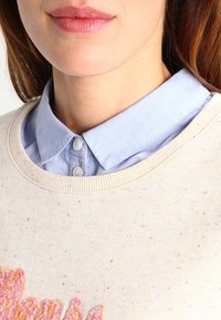 Pull beige clair avec des éclats colorés, doté d'un col rond et d'un motif floral texturé rose. En dessous, une chemise à bouton bleu clair.