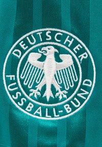 Κεντητό λευκό έμβλημα αετού με το κείμενο "Deutscher Fussball-Bund" σε φόντο πράσινου ριγωτού υφάσματος.