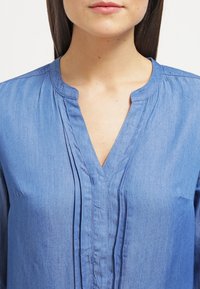 Jasnoniebieska bluzka z chambray o dekolcie w kształcie litery V, plisowanym przodzie i zaokrąglonym dekolcie. Gładka faktura, długie rękawy i minimalne detale.