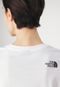 Bílý bavlněný tričko s kulatým výstřihem. Na zadní straně se nachází černé logo "The North Face" v levém horním rohu.