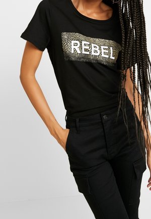 Czarny bawełniany t-shirt z złoto-czarnym wzorem w kształcie prostokąta przedstawiającym napis "REBEL" na białym tle. Połączony z czarnymi spodniami, widok z boku.