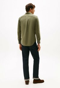 Camisa de manga larga verde oliva con corte recto, puños abotonados y pliegue trasero cosido, combinada con jeans azul oscuro y zapatos marrones.