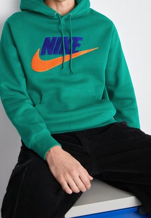Osoba siedząca w zielonej bluzie z kapturem z dużym pomarańczowym znakiem Nike oraz niebieskim napisem "NIKE", w połączeniu z czarnymi sztruksowymi spodniami.