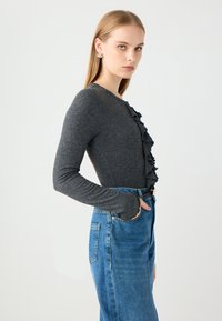 Grijze lange mouwen top met ruches aan de voorkant, gecombineerd met hooggeplaatste blauwe spijkerbroeken. De outfit heeft een aansluitende silhoeëtten en een geweven stof.