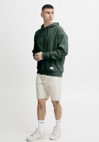 Sudadera verde con capucha y cordones, puños acanalados y un bolsillo frontal. Combinada con pantalones cortos beige y zapatillas blancas.