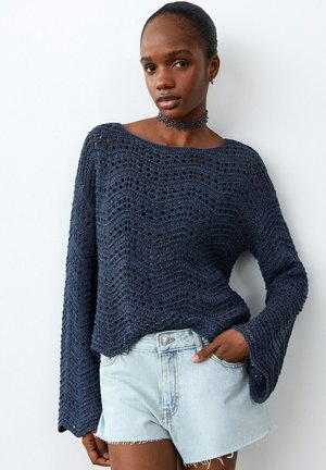 Femme portant un pull en maille ouverte bleu marine avec des manches cloche et un short en jean clair découpé, debout devant un fond blanc.