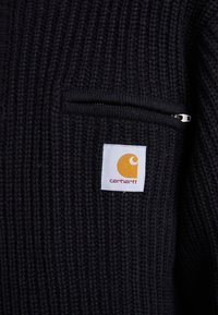Črn pleten pulover z zadrgo in kvadratno nalepko z oranžnim logotipom in besedilom "carhartt". Teksturovani rebrasti material.