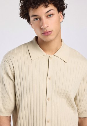 Jong persoon met krullend haar die een beige geribd kort mouw knoopshirt draagt en lichtjes schuin naar de camera kijkt.