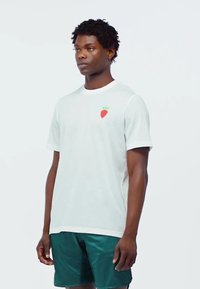 Camiseta de algodón blanca con mangas cortas; presenta un pequeño gráfico de una fresa roja en el lado izquierdo del pecho. Se combina con pantalones cortos de color verde oscuro.