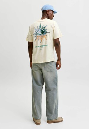 Mann trägt cremefarbenes T-Shirt mit Sonne- und Landschaftsaufdruck auf dem Rücken, hellblaue Mütze, lockere, ausgebleichte Jeans und beige Schuhe, seitlich stehend.