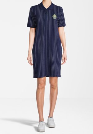 Mujer que lleva un vestido polo azul marino hasta la rodilla con rayas verticales blancas y un escudo en el pecho, combinado con zapatillas blancas informales.