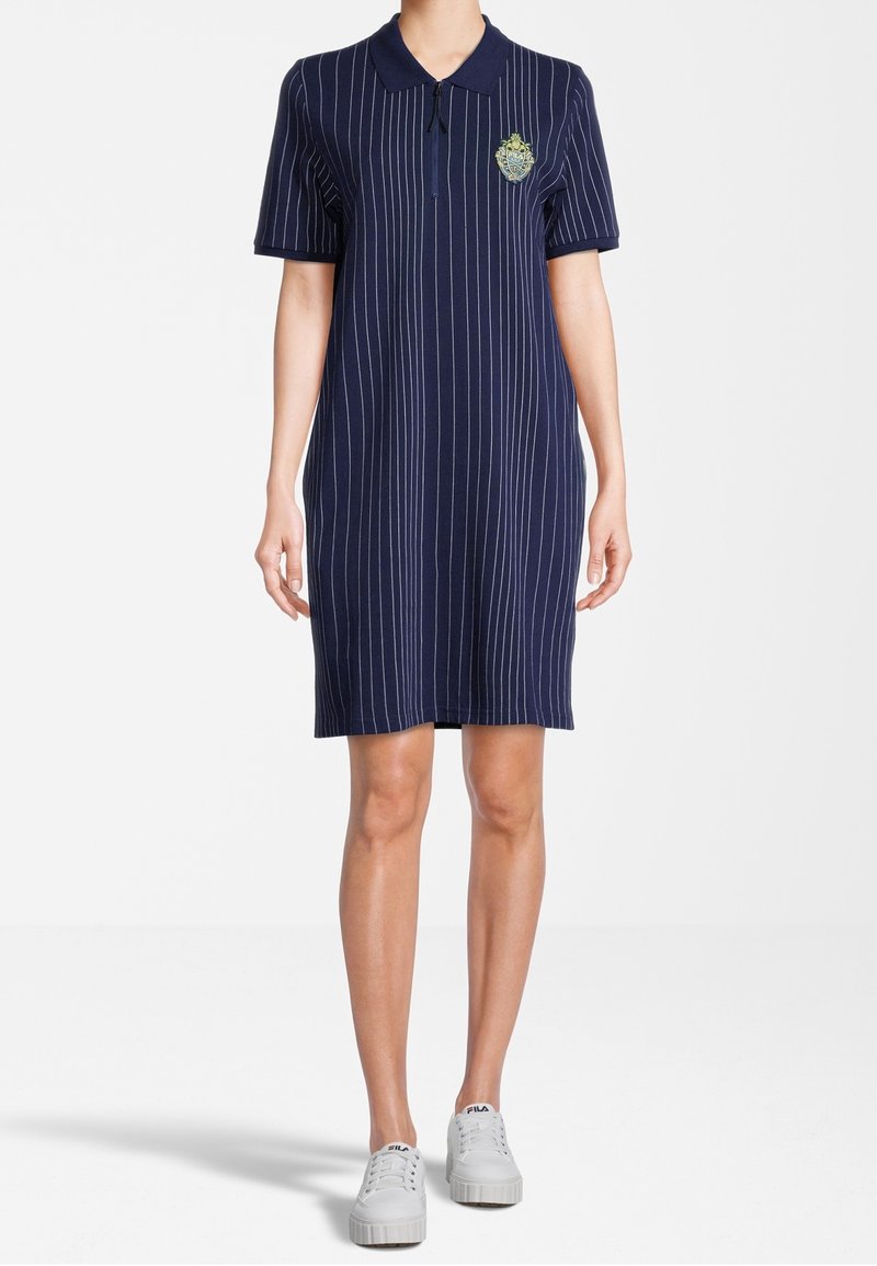 Femme portant une robe polo bleu marine longueur genou avec des rayures verticales blanches et un blason sur la poitrine, associée à des baskets blanches décontractées.