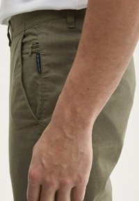 Gros plan sur la main d'une personne reposant à côté d'un pantalon vert olive avec une petite poche zippée et une étiquette de marque en tissu visible.