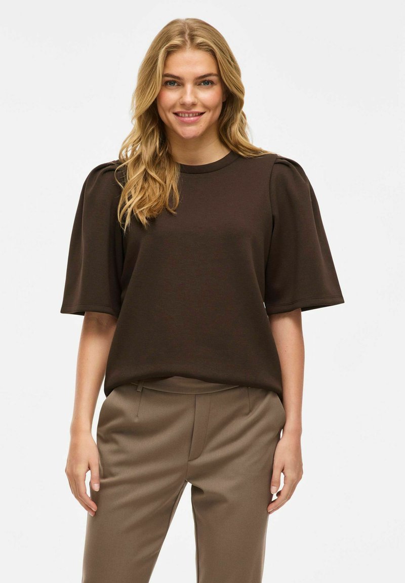 Brūns īsām piedurknēm tops ar noapaļotu kakla izmēru un puffed pleciem, izgatavots no gludas auduma. Apvienots ar pielāgotiem beigu atliktiem biksēm.