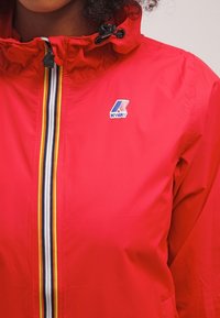 Czerwona wodoodporna kurtka z wysokim kołnierzem, z centralnym zamkiem błyskawicznym i wielokolorowym pionowym paskiem. Logo K-Way na górze po lewej stronie.