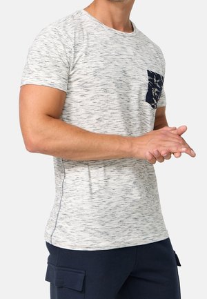 Mand iført en lysegrå t-shirt med mørkt mønstret lomme og marineblå cargobukser, hænderne foldet foran.