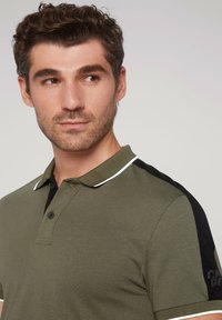 Olivgrünes Poloshirt mit schwarzem Kragen und Ärmeln, weißen Paspeln und einer Knopfleiste mit zwei Knöpfen. Weicher Stoff mit schlanker Passform.