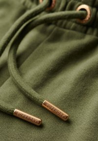 Tela verde con una superficie texturizada, que incluye cordones con puntas color cobre grabadas con la palabra "SUPERDRY" para añadir detalles.