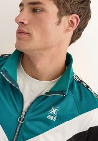 Teal zippered jacka med vita och svarta detaljer, med en logotyp på bröstet, mjukt tyg och en hög krage.