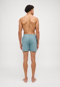 Lyseblå badebukser laget av et glatt, hurtig tørkende materiale, har elastisk midje og en baklomme.