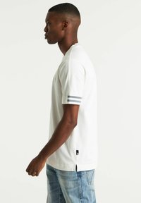 T-shirt bianco in cotone con maniche corte e strisce grigie. Include spacchi laterali e un piccolo etichetta del marchio sul lato sinistro.