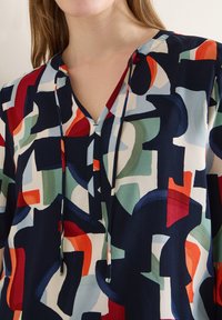 Femme portant un chemisier multicolore avec des formes géométriques abstraites en bleu marine, rouge, vert et blanc, doté d'un col en V et de liens à nouer.