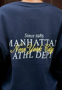 Tummansininen collegepaita, jonka selässä on valkoisella ja keltaisella tekstillä kirjoitettu "Since 1985 Manhattan New York City ATHL DEPT".