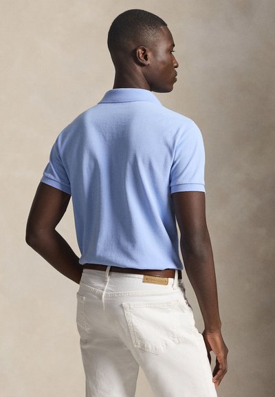 Άνδρας που φοράει ανοιχτό μπλε κοντομάνικο πόλο και άσπρο παντελόνι, στέκεται με το ένα χέρι στη μέση, στραμμένος προς τα πίσω πάνω σε ουδέτερο φόντο.