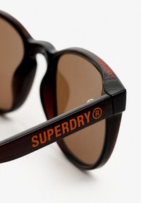 Gafas de sol de tono marrón con una forma clásica de gran tamaño. Montura negra y de carey con un destacado logo naranja "SUPERDRY" en las patillas.