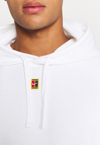 Gros plan d'une personne portant un sweat à capuche blanc avec un patch du logo Nike multicolore sur la poitrine et des cordons blancs.