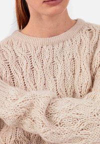 Pull beige clair en tricot avec un motif torsadé texturé, un col large côtelé et des manches larges et confortables. Matière douce et épaisse en tricot.
