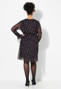 Schwarzes Kleid mit langen transparenten Ärmeln, mit einem roten und weißen Herzmuster, tailliert und knieumspielend. Schwarze Strumpfhosen und Absatzschuhe.