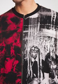 Black and red zip-up jacket featuring graphic prints of figures with masks, contrasting textures, and a round neckline.