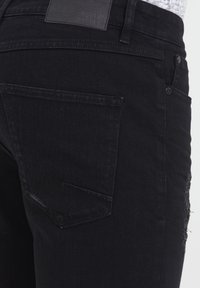 Mustad teksapüksid, millel on kulunud osa ja tagatasku diagonaalse kujundusega. Vöökohal on brändinimetusega nahkne märk.