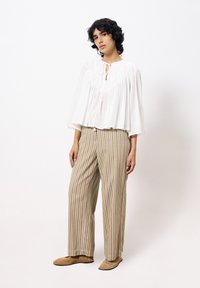 Weiße, strukturierte Bluse mit langen Ärmeln und Bindegurt, kombiniert mit beigen, gestreiften, weiten Hosen. Modell trägt beige Hausschuhe.