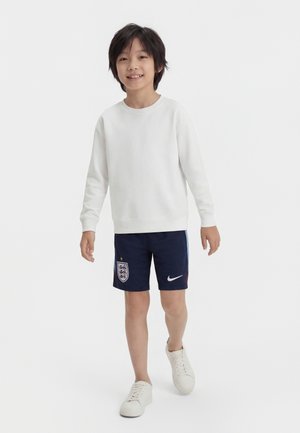 Ung dreng gående iført en hvid langærmet skjorte, marineblå England fodboldshorts med logoer og hvide sneakers, på en ensfarvet baggrund.