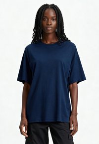 Kleita zila, pārāk liela kokvilnas t-krekls ar īsām piedurknēm un apaļu kakla izgriezumu, attēlota uz gaiša fona. Gluda tekstūra.