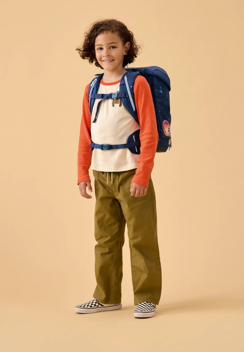 Sac à dos bleu avec motif étoilé, chemise à manches longues orange, tee-shirt beige, pantalon vert et chaussures à damier noir et blanc.