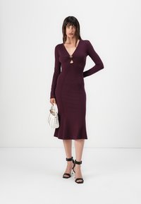 Lila Strickkleid mit langen Ärmeln, figurbetontem Schnitt, Ausschnitt mit Schlitz und ausgestellt geschnittenem Saum. Schwarze Riemchen-Heels und weiße Tasche.