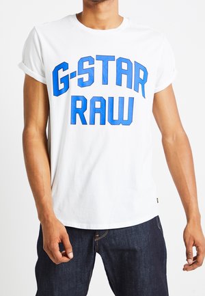Homme portant un t-shirt blanc à manches courtes avec des revers roulés et un texte bleu "G-STAR RAW", associé à un jean en denim foncé.