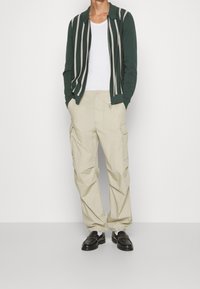 Grön zip-up kofta med kontrasterande beige ränder, vit t-shirt, beige cargobyxor och svarta loafers. Enkelt, avslappnat snitt.