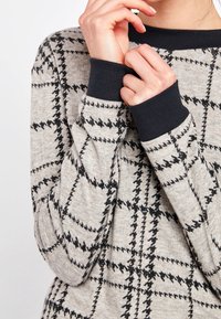 Haut beige à manches longues avec un motif géométrique noir, poignets et col en maille côtelée bleu marine, texture douce, présentant un gros plan des manches.