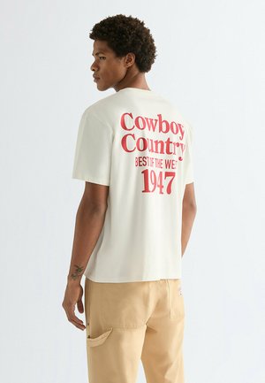 Giovane uomo che indossa una maglietta color crema con testo rosso "Cowboy Country Best of the West 1947" e pantaloni beige, in piedi di lato rivolto verso sinistra.