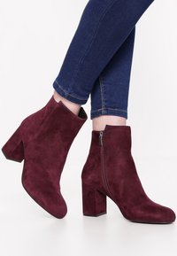Personne portant des bottines en daim bourgogne avec des talons carrés et des fermetures éclair sur le côté, associées à un jean skinny bleu foncé.