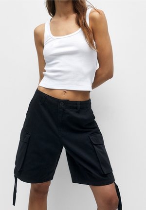 Kvinde iført en hvid cropped tanktop og sort cargo-shorts til knæene med lommer foran, stående mod en ensfarvet baggrund.