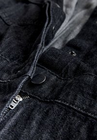 Gros plan sur un tissu denim noir avec une fermeture éclair en métal visible et des boutons-pression sur un vêtement.