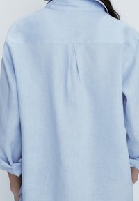 Camicetta azzurra chiara a maniche lunghe realizzata in un tessuto morbido. Presenta un colletto, una piega sul retro e maniche arrotolate. La texture appare liscia e leggera.