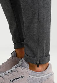 Grå texturerade joggers med detaljerade dragkedjor vid ankeln, tillsammans med ljusgrå Reebok-sneakers med logotyp och snörningsdesign.