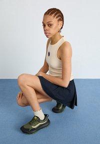 Haut beige sans manches, jupe bleu marine et baskets vert olive avec des accents noirs et jaunes. Cheveux courts avec des tresses et des boucles d'oreilles.